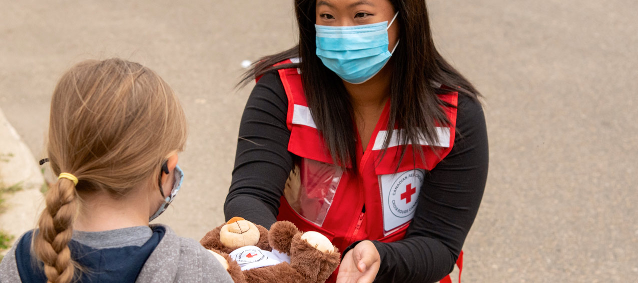 A Red Cross volunteer wearing a face masks hands a teddy bear to a young girl.
