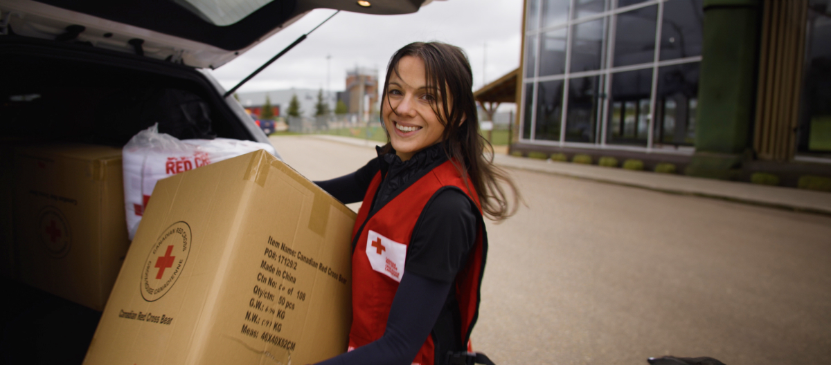 A Red Cross staff member carries a box of supplies and smiles at the camera.