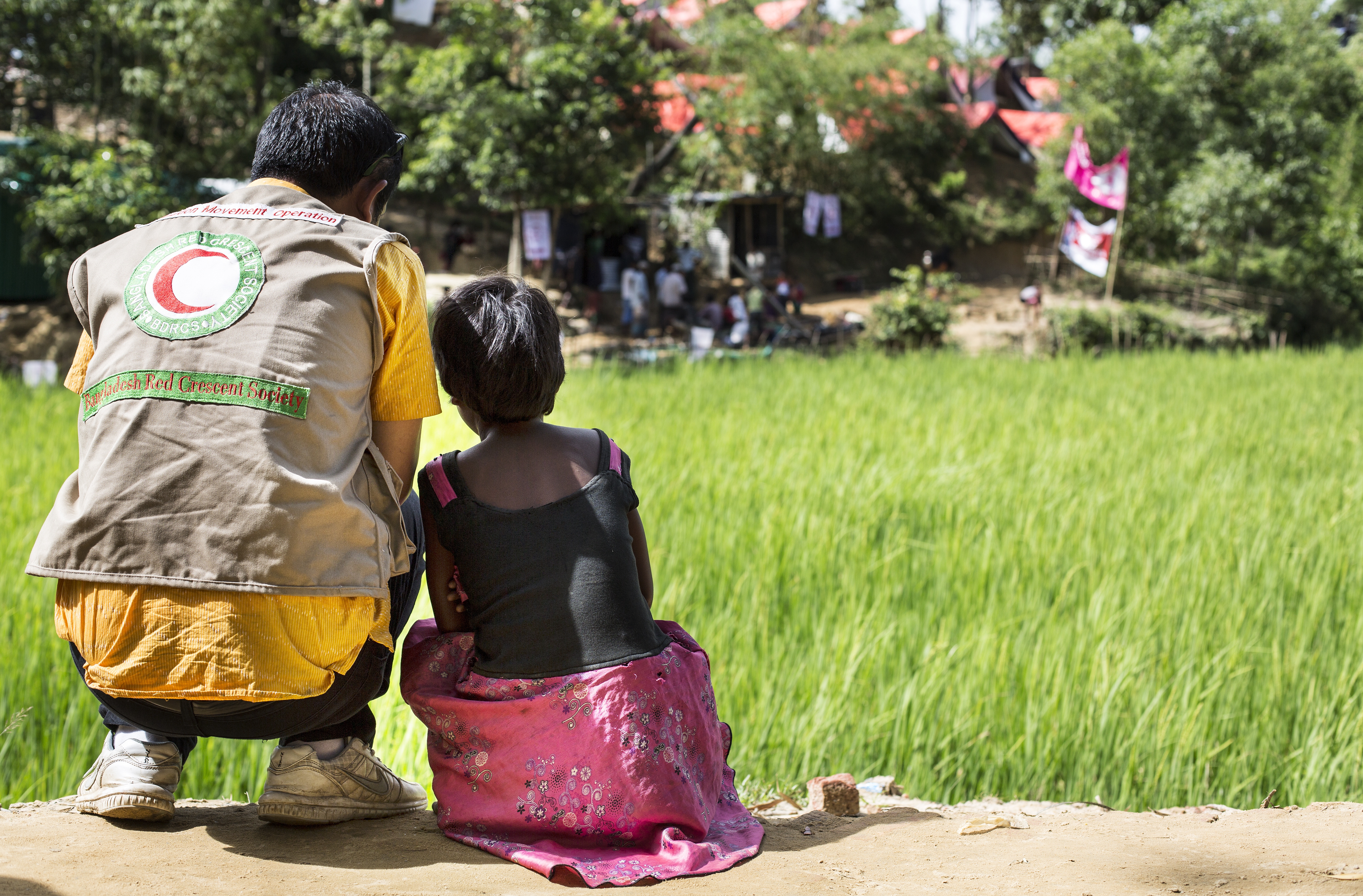 A man and a child sitting together on the ground, looking ahead at a vast green field