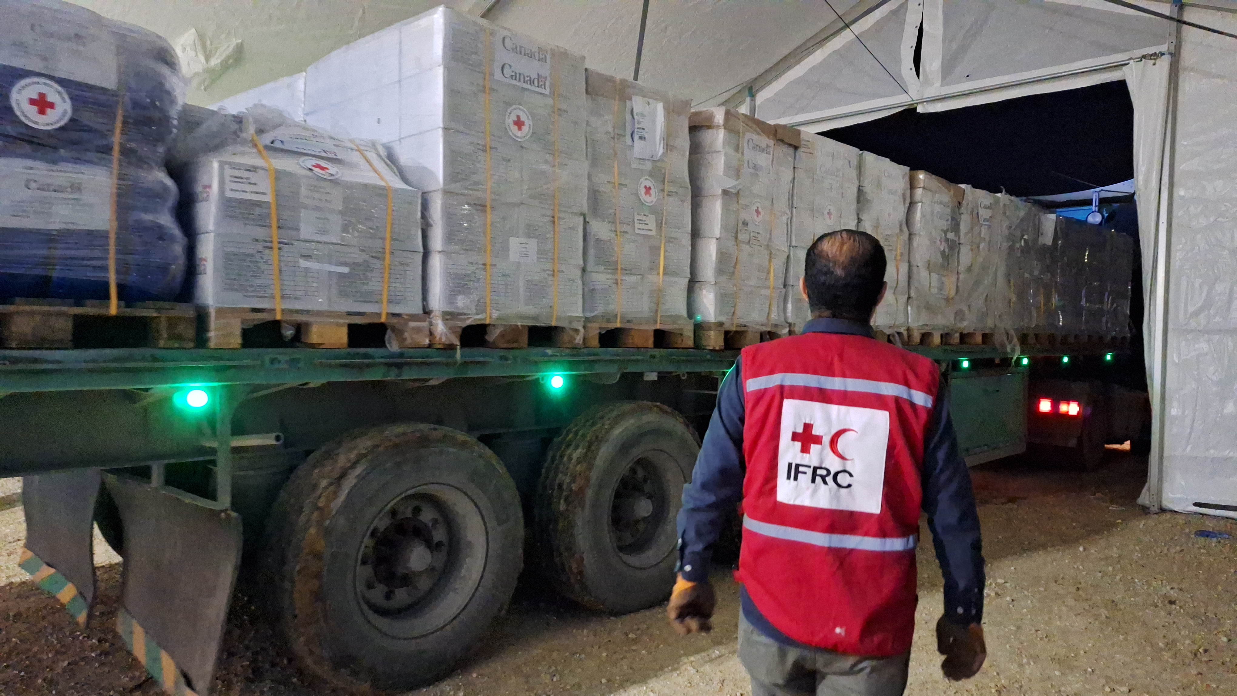 A man in a red IFRC vest stands in front of a truck with humanitarian supplies.