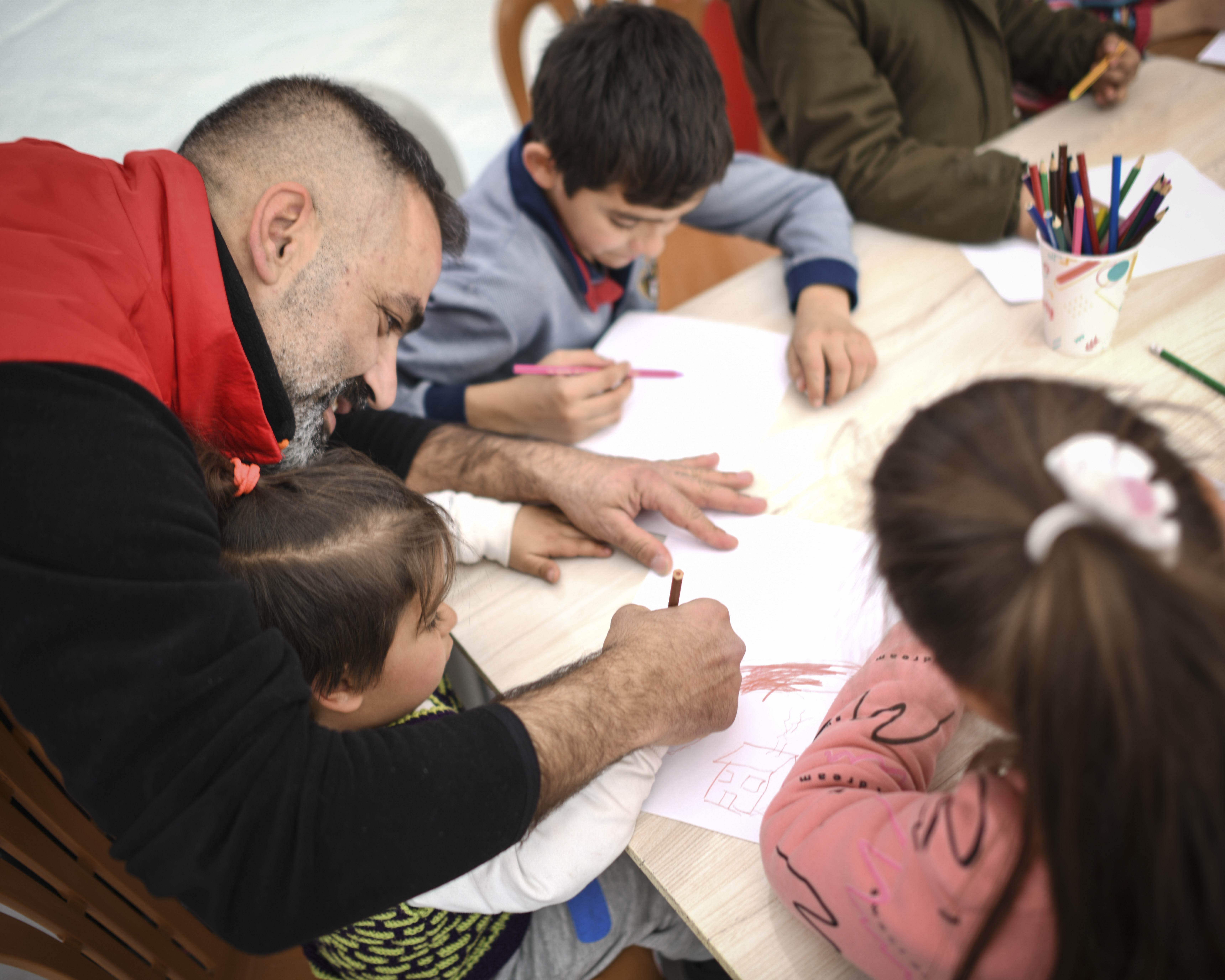 Three children sit at a table drawing while a volunteer in a red vest helps.