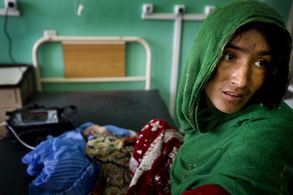A worried looking mother sits on a hospital bed which her infant is resting in