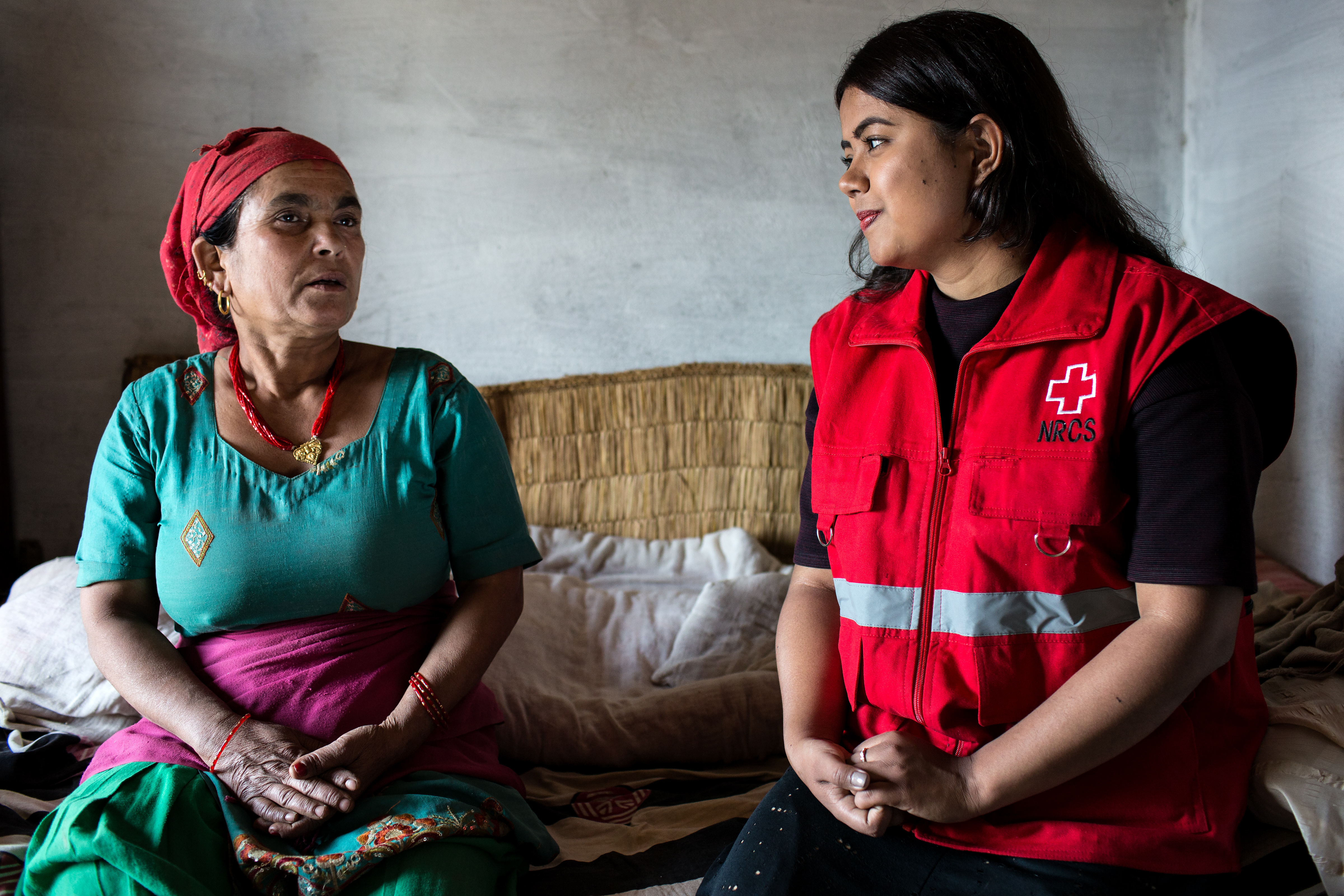 Two women sat on a bed talking with one woman wearing a Red Cross vest