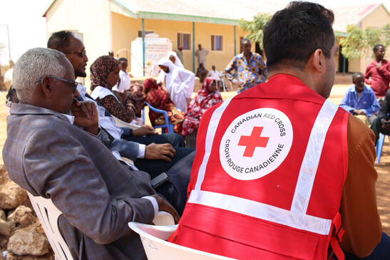 Group of people sitting in a circle outside with one wearing a Red Cross vest