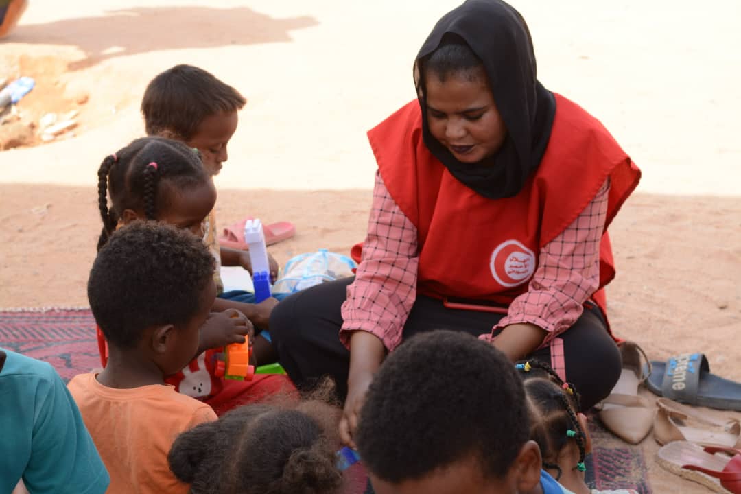 A woman in a red vest sitting on the ground outdoors playing with children