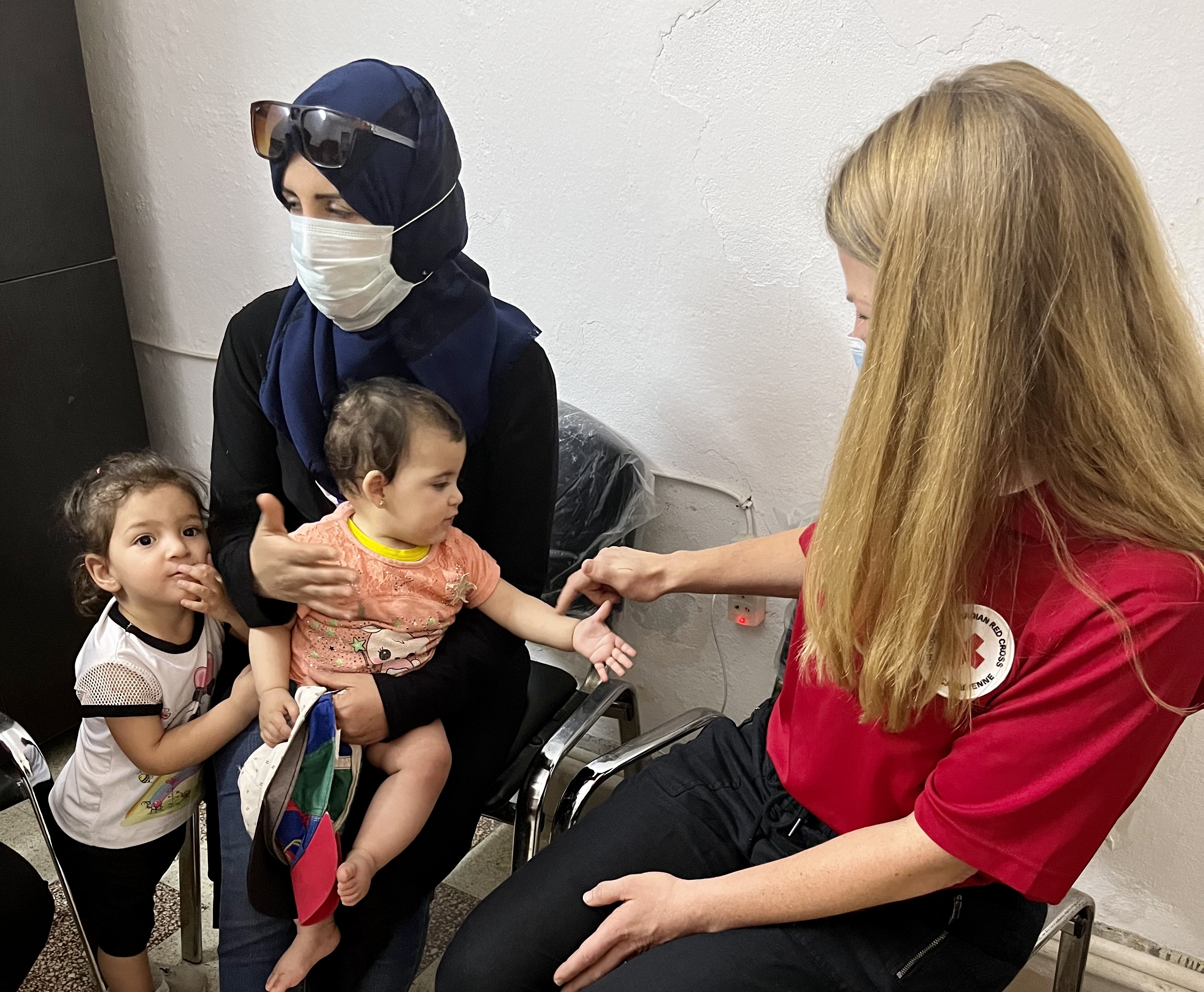 A seated woman holding a baby is seated beside a child and woman in a Red Cross shirt